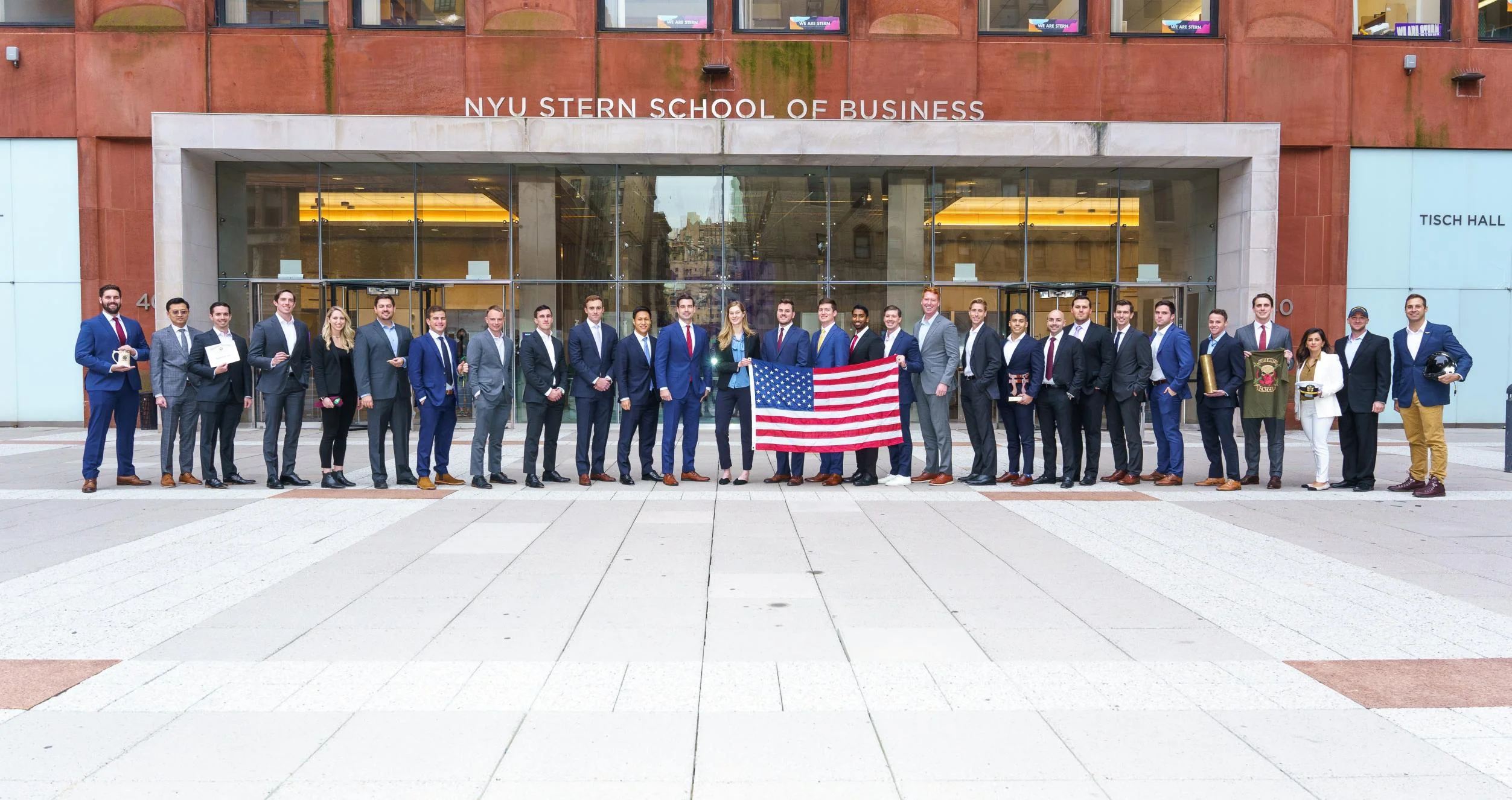 A large line of NYU veterans in formal wear holding an American flag outside the Stern School of Business